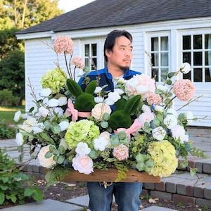 May include: A wooden box overflowing with an arrangement of spring flowers and greenery. Two green moss-covered bunny heads with pink ribbons are nestled among the blooms. The arrangement includes white, pink, and yellow flowers.