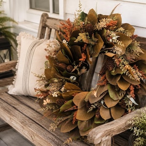 May include: A circular wreath featuring brown magnolia leaves, small pinecones, and textured orange and green foliage. The arrangement is composed of layered botanical elements in earthy tones, creating a dense, rounded display of natural textures.