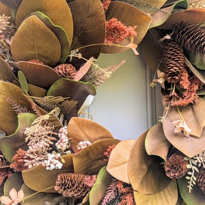 May include: A circular wreath featuring large brown and green magnolia leaves, interspersed with brown pinecones, textured rust-colored foliage, and small clusters of pale pink berries. The arrangement creates a dense, layered botanical display.
