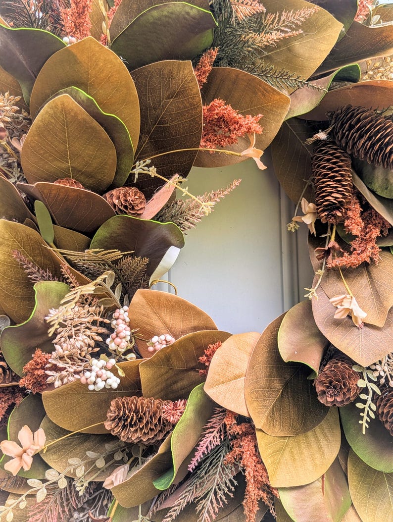 May include: A circular wreath featuring large brown and green magnolia leaves, interspersed with brown pinecones, textured rust-colored foliage, and small clusters of pale pink berries. The arrangement creates a dense, layered botanical display.