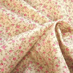 May include: A close-up of a cream-coloured fabric with a delicate floral pattern. Small pink roses and green leaves are scattered across the material. The fabric is folded, showing the texture and drape of the material.