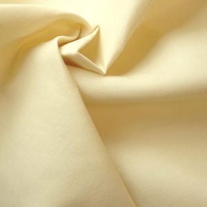 May include: A close-up shot of a folded piece of pale yellow fabric. The fabric has a smooth texture with subtle woven lines. The folds create shadows and highlights, adding depth. The overall impression is of a soft, natural material, possibly cotton or linen.