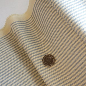May include: Close-up of a cream-coloured fabric with thin blue stripes. A British coin is placed on the fabric for scale.