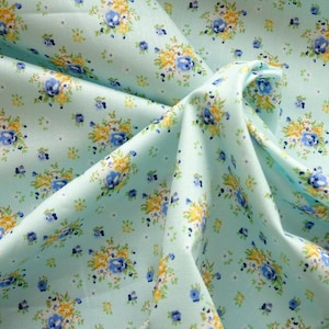 May include: Close-up of a pale blue fabric with a floral design. The pattern includes clusters of yellow and blue flowers with green leaves. The fabric is folded, revealing the texture and detail of the floral arrangement. The overall effect is delicate.