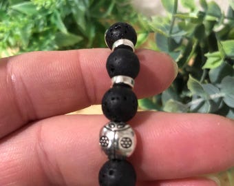 May include: A black lava stone bracelet with silver-colored accents. The bracelet features round, porous black beads separated by silver-toned spacers. One bead has a decorative silver design. The bracelet is held in a hand.