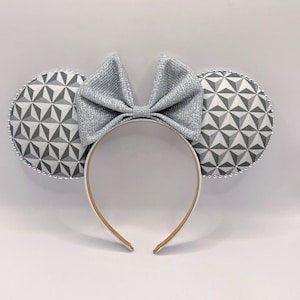 May include: A headband featuring two circular ears with a gray and white geometric triangular pattern and a silver glittery bow centered between them. The edges of the ears are lined with small silver beads, and the headband has a plain white band.