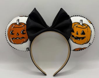 May include: A Halloween headband with two white circular ear accents, each displaying an orange jack-o'-lantern design. A large black bow is positioned above the headband, which has a tan interior and black exterior. The ears are trimmed with black beading.