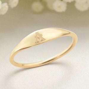 Puede incluir: Un anillo dorado con una parte superior lisa en forma ovalada. El anillo presenta un delicado grabado de la letra "A" en la parte superior. El anillo se encuentra sobre un fondo suave y neutro, con flores blancas en la esquina superior izquierda.