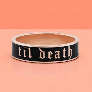May include: A gold-toned ring with a black enamel band. The phrase "til death" is inscribed in a gothic font around the band. The ring is set against a peach and pink background.