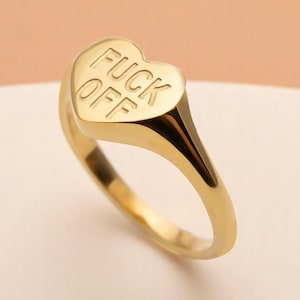 May include: A gold-colored signet ring with a heart-shaped face. The ring's face has the words "FUCK OFF" engraved in capital letters. The ring is made of a shiny, reflective metal.