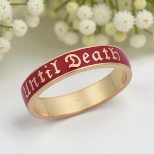 May include: Gold-tone ring with a red enamel band. The ring features the inscription "until Death" in an elegant, stylized font. The ring is set against a soft, blurred background of white flowers, highlighting the jewelry's design.