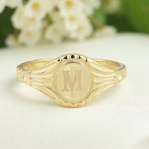 May include: A gold-colored signet ring with the letter "M" engraved in the center. The ring has a rounded oval face and a detailed band. The ring is displayed on a white surface.