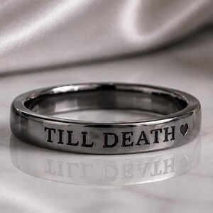 May include: A polished gunmetal gray band ring featuring the engraved text TILL DEATH followed by a small heart symbol. The ring has a smooth, reflective metallic finish and a simple, rounded profile, showcasing clear, dark lettering on the outer surface.