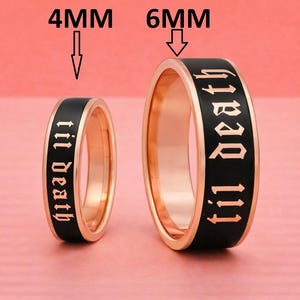 以下が含まれることがあります： 黒いバンドが付いた2つの金色の指輪。指輪にはゴシック体で「til death」というフレーズが書かれています。1つのリングは幅4mm、もう1つは6mmです。リングはピンク色の背景に表示されています。