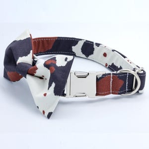 May include: A white, brown, and black patterned dog collar with a silver buckle and a matching bow tie.