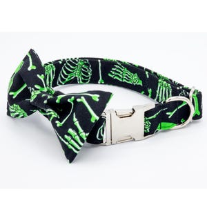May include: A black dog collar with a green skeleton print and a matching bow tie. The collar has a silver buckle and a silver D-ring.