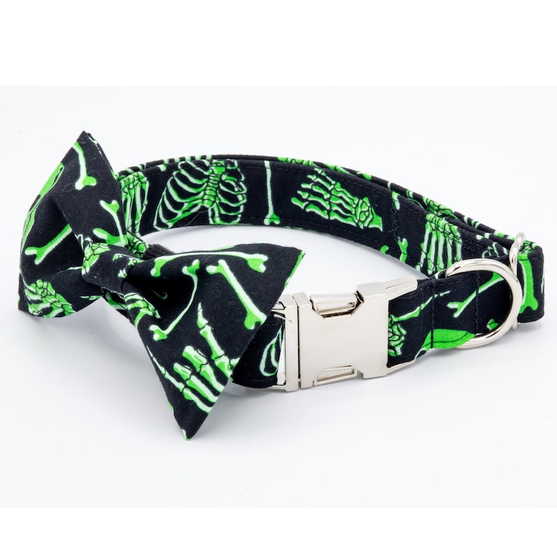 May include: A black dog collar with a green skeleton print and a matching bow tie. The collar has a silver buckle and a silver D-ring.