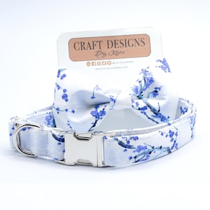 May include: White dog collar and matching bow tie with a blue floral pattern. The collar has a silver buckle and a silver D-ring. The bow tie is tied with a white ribbon. The collar and bow tie are both made of fabric.