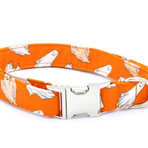 May include: Orange dog collar with a repeating pattern of white cartoon ghosts. The collar has a silver metal buckle and a D-ring for leash attachment. The collar is designed for pets.