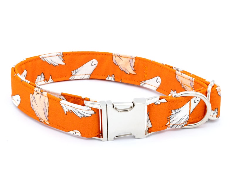May include: Orange dog collar with a repeating pattern of white cartoon ghosts. The collar has a silver metal buckle and a D-ring for leash attachment. The collar is designed for pets.
