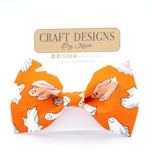 May include: Orange bow tie with a repeating pattern of white cartoon ghosts. The bow tie is centered on a white background. A brown tag with the text "Craft Designs By Kenz" is above the bow tie.