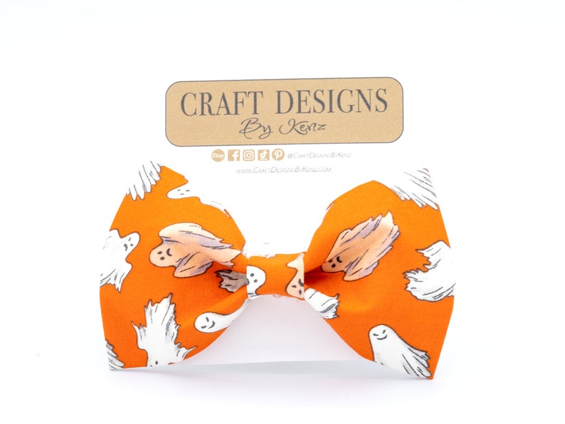 May include: Orange bow tie with a repeating pattern of white cartoon ghosts. The bow tie is centered on a white background. A brown tag with the text "Craft Designs By Kenz" is above the bow tie.
