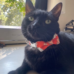 May include: A black cat wearing a Halloween-themed bow tie collar. The bow tie is orange with a white ghost pattern. The cat has green eyes and is looking towards the camera. The cat is lying down on a white surface.
