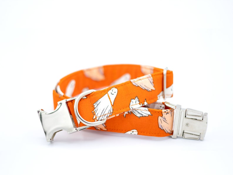 May include: An orange dog collar with a white ghost pattern. The collar has a silver metal buckle and D-ring. The fabric is a bright orange with white ghost illustrations. The collar is designed for pets.