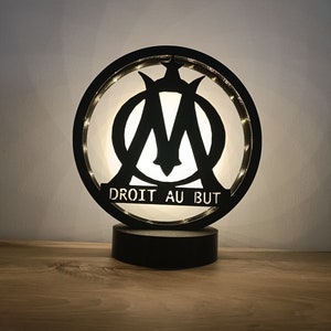 Könnte beinhalten: Eine schwarze und weiße Holzlampe mit einem runden Design, das den Buchstaben "M" in einem Kreis mit den Worten "DROIT AU BUT" darunter zeigt. Die Lampe ist beleuchtet und strahlt ein warmes Licht aus.