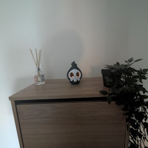 Puede incluir: Un portavelas decorativo negro con forma de calavera con ojos naranjas y una capucha negra está sobre un mueble de madera. Una botella difusora de vidrio con cañas y una pequeña planta en maceta también están en el mueble. El mueble tiene un acabado marrón oscuro.