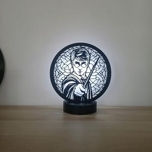 Może przedstawiać: Czarno-biała sylwetka Harry'ego Pottera trzymającego różdżkę, z witrażowym wzorem w tle. Sylwetka stoi na czarnej okrągłej podstawie, przez którą świeci białe światło.