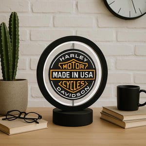 Può includere: Un'insegna al neon nera Harley-Davidson con un contorno al neon bianco. L'insegna presenta il logo Harley-Davidson con la scritta "MADE IN USA". L'insegna è su una base nera, con un cactus e una tazza nera sullo sfondo.