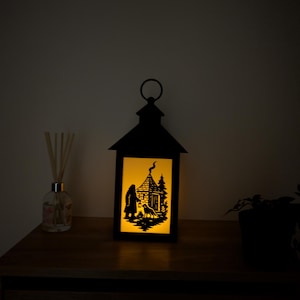 Op de afbeelding: Zwarte metalen lantaarn met een silhouetscène van een huis, persoon en hond, verlicht door warm geel licht. De lantaarn heeft een handvat en staat op een houten oppervlak.