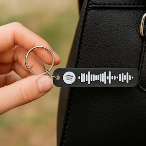 Può includere: Un portachiavi nero con codice Spotify e anello argentato. Il portachiavi è tenuto davanti a una borsa in pelle nera. Il logo Spotify e un motivo a onde sonore sono visibili sul portachiavi.