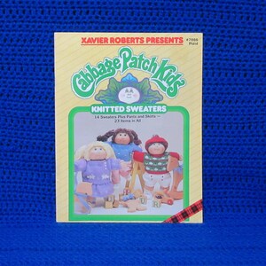 May include: A vintage Cabbage Patch Kids book cover featuring the text "KNITTED SWEATERS." The cover has a green and yellow border with illustrations of Cabbage Patch Kids dolls. The background is a vibrant blue.
