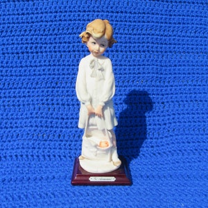 May include: A porcelain figurine of a young girl in a white dress holding a basket with a baby inside. The figurine is on a wooden base with the name "G. Armani" inscribed on it.
