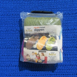 May include: A plastic package containing a crochet kit for making slippers. The package shows a family wearing the slippers and the text "minute crochet slippers family favorites...fast 'n easy to make" and "All you need to make 1 pair of slippers".