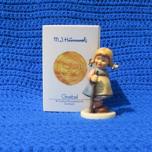 May include: A porcelain figurine of a girl with pigtails holding a stick. She is wearing a blue and white dress and a red jacket. The figurine is on a white base and is part of a Goebel collection. The box is white with the Goebel logo and the words "M.J. Hummel" in gold.