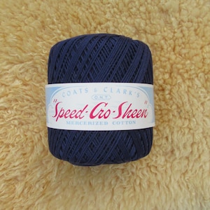 May include: A dark blue spool of Coats & Clark's Speed-Cro-Sheen mercerized cotton yarn. The yarn is wrapped with a white label featuring the brand name in red script. The background is a textured, light tan surface.