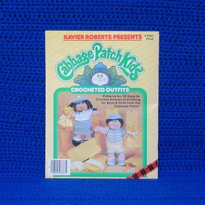 May include: A vintage Cabbage Patch Kids crochet pattern book. The cover features the text "XAVIER ROBERTS PRESENTS Cabbage Patch Kids CROCHETED OUTFITS". The book shows two dolls wearing crocheted outfits. The background is a vibrant blue.