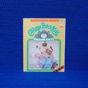 May include: A vintage Cabbage Patch Kids Preemie Knits pattern book. The cover features the Cabbage Patch Kids logo, dolls, and text. The book includes patterns for blankets, sleepers, booties, bonnets, and more. The background is a vibrant blue.
