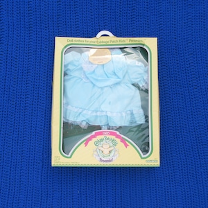 May include: A boxed set of doll clothes for Cabbage Patch Kids Preemies. The light blue dress has white lace trim and is visible through the clear plastic window. The box has a yellow and green design with the Cabbage Patch Kids logo and the year 1986.