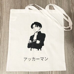 以下が含まれることがあります： 白地のキャンバス地のトートバッグに、黒と白のアニメキャラクターのイラストと日本語で「進撃の巨人」の文字が書かれている。