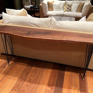 May include: A long, dark wood console table with a live edge top and black hairpin legs. The table is in front of a beige sofa with white pillows.