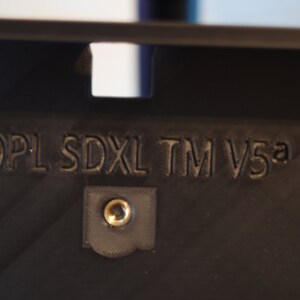 May include: Close-up of a black plastic device with the text "4DPL SDXL TM V5a" printed on it. There is a small screw in the center of the device.