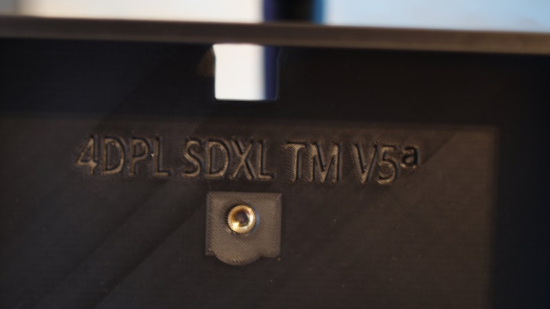 May include: Close-up of a black plastic device with the text "4DPL SDXL TM V5a" printed on it. There is a small screw in the center of the device.