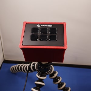 May include: A black Stream Deck with eight buttons, encased in a red silicone case, mounted on a black and white tripod.