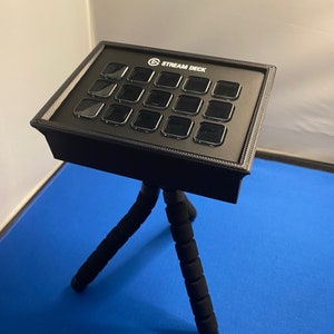 Puede incluir: Un Stream Deck negro con 15 botones está montado en un trípode negro. El trípode tiene tres patas con acolchado de espuma negra.