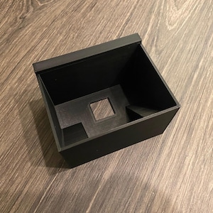 May include: A black plastic box with a square opening in the top and a smaller rectangular opening on the side. The box is designed to hold small items.