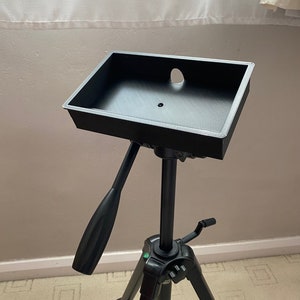 May include: Black tripod with a black tray on top. The tray has a hole in the center. The tripod has a green button on the side. The tripod is made by Velbon.